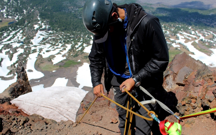 Wearing safety gear and secured by ropes, a person stands on the edge of a cliff, high above a landscape dotted with snow.